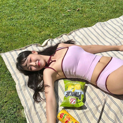 Women's light purple one-shoulder swimsuit with dark red contrast trim, lying on a striped blanket on green grass. - Image 4