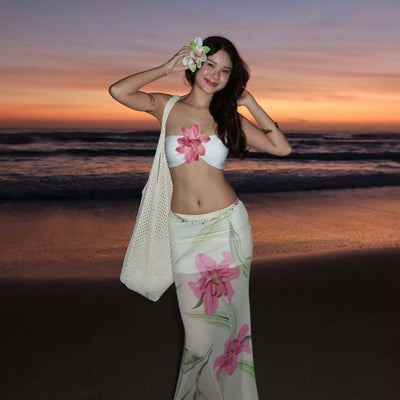 Woman in a white tube top with pink flower and a sheer floral maxi skirt, posing on a beach at sunset. - Image 6
