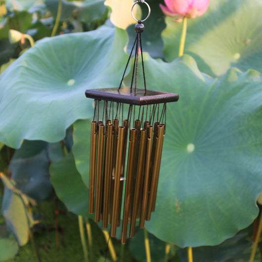 Bronze wind chime with multiple metal tubes hanging from a dark rectangular top, set against green lotus leaves. - Image 11