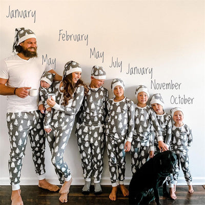 Family of seven and a dog in matching grey Christmas tree print pajamas with white cuffs and beanies.
