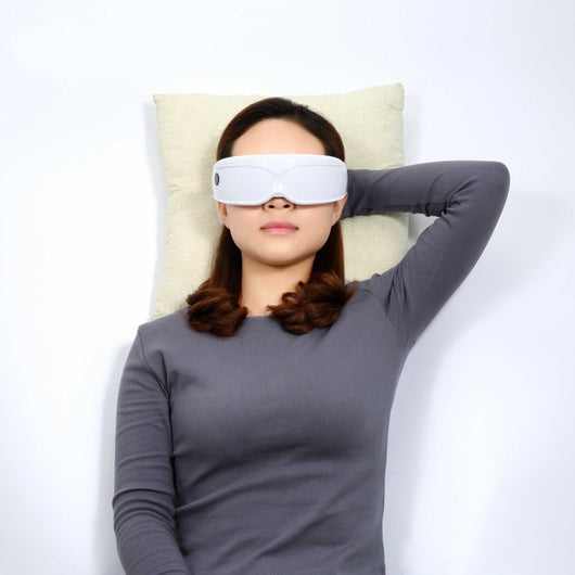 White and black eye massager with a goggle-like design, featuring a white frame and dark lenses, on a wooden surface. - Image 2