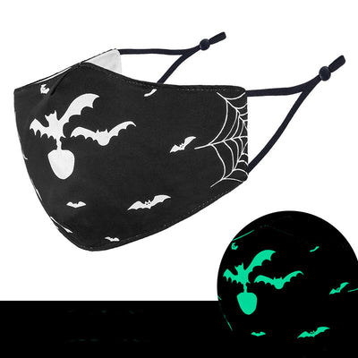Black cloth face mask with white Halloween witch, bats, and jack-o'-lantern pattern, shown glowing green in the dark.