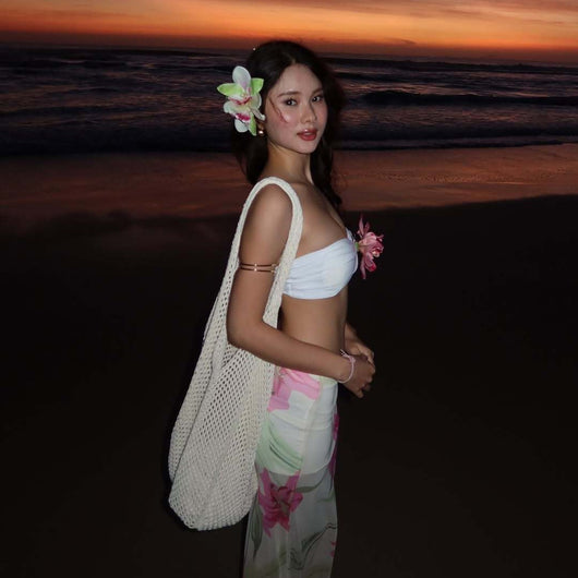 Woman in a white tube top with pink flower and a sheer floral maxi skirt, posing on a beach at sunset. - Image 2