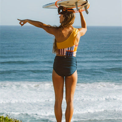 Smiling woman in a one-shoulder, color-block bikini top and high-waisted bottom on a sandy beach. - Image 9