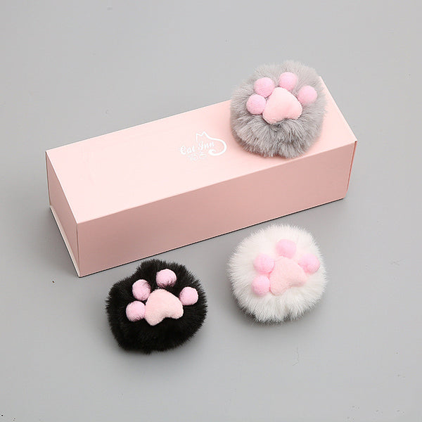 Three fluffy cat toy balls in white with pink paw print, gray, and black, next to a pink gift box.