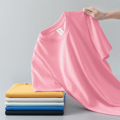 Light gray short-sleeve t-shirt with a round neck, held up to show its smooth fabric. A stack of folded t-shirts in various colors is visible in the background. - Image 6