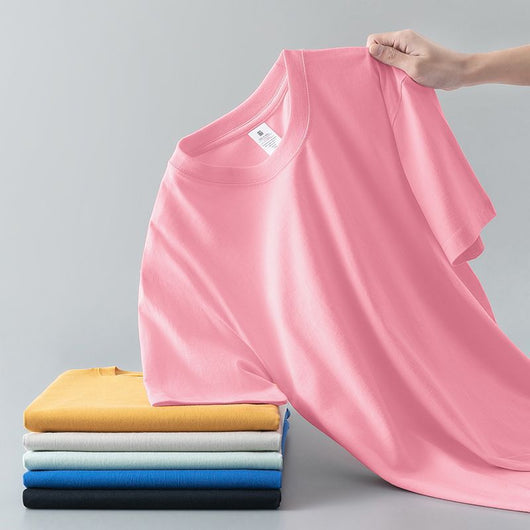 Light gray short-sleeve t-shirt with a round neck, held up to show its smooth fabric. A stack of folded t-shirts in various colors is visible in the background. - Image 6