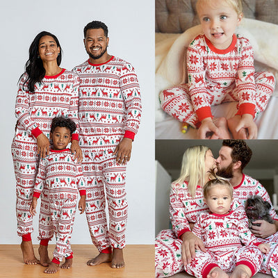 Family wearing matching white and red Christmas pajamas with elk and snowflake patterns, including a baby and a dog.