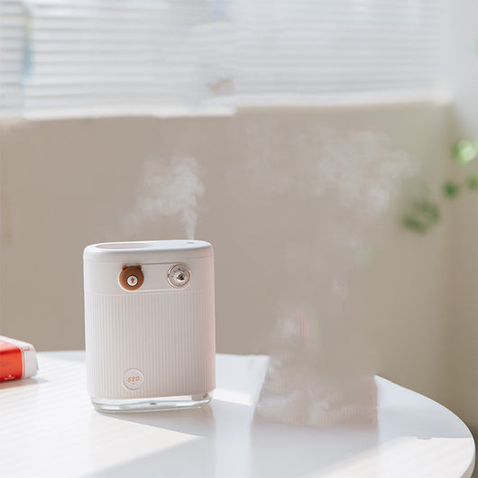 White cylindrical humidifier with ribbed texture, clear base, brown bear button, and silver dial, emitting mist on a white table. - Image 6