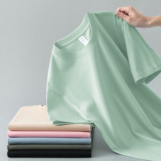 Light gray short-sleeve t-shirt with a round neck, held up to show its smooth fabric. A stack of folded t-shirts in various colors is visible in the background.