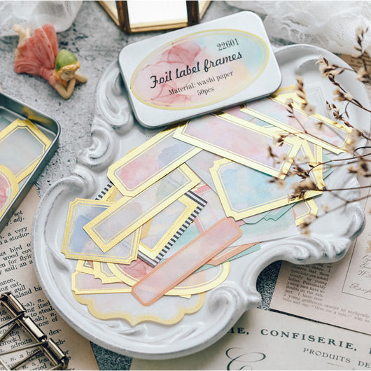 Assorted decorative paper labels with gold foil borders and soft watercolor designs in pink, blue, and green hues.
