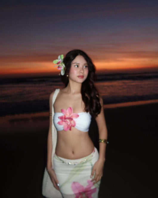 Woman in a white tube top with pink flower and a sheer floral maxi skirt, posing on a beach at sunset. - Image 11