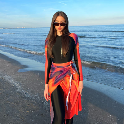 Woman in a black long-sleeve rash guard, black swim leggings, and a vibrant tie-dye sarong on a sunny beach. - Image 2