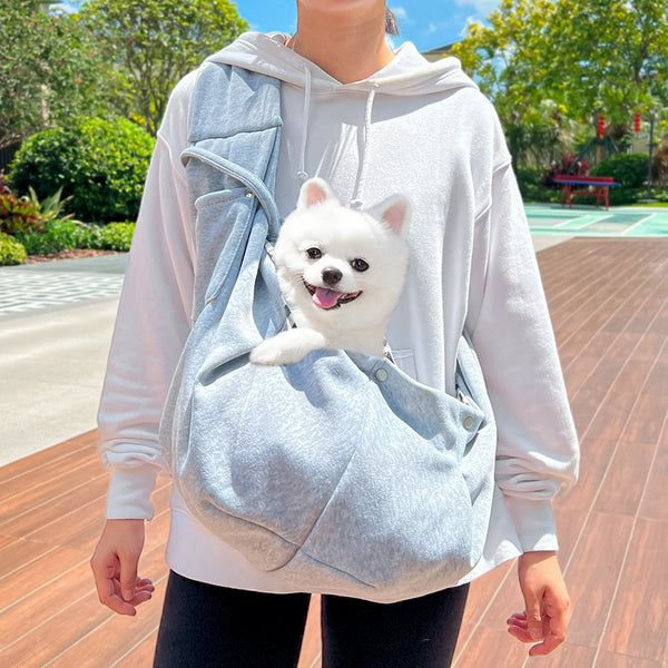 Person in white hoodie carrying a happy white dog in a navy blue pet sling bag outdoors.
