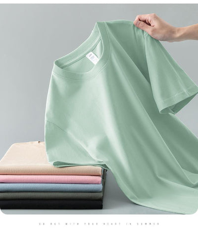 Light gray short-sleeve t-shirt with a round neck, held up to show its smooth fabric. A stack of folded t-shirts in various colors is visible in the background. - Image 19