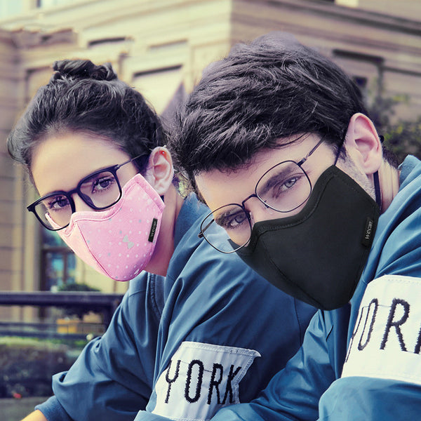 Woman wearing a pink cotton face mask with light blue polka dots and bow patterns, paired with black-framed glasses.