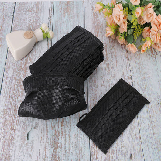 Stack of black disposable 3-ply face masks with ear loops, and one mask laid flat on a light wood surface.