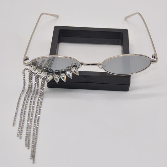 Silver oval-frame sunglasses with mirrored lenses and glamorous rhinestone tassels on one side, held by a gloved hand.