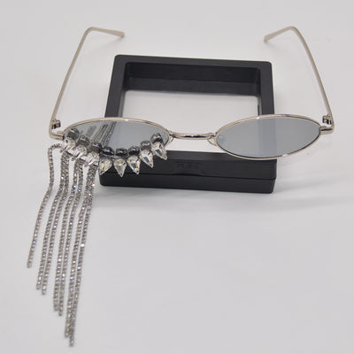 Silver oval-frame sunglasses with mirrored lenses and glamorous rhinestone tassels on one side, held by a gloved hand.