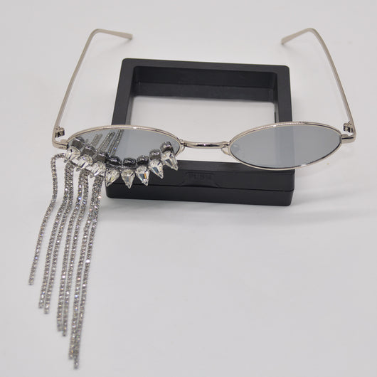Silver oval-frame sunglasses with mirrored lenses and glamorous rhinestone tassels on one side, held by a gloved hand.