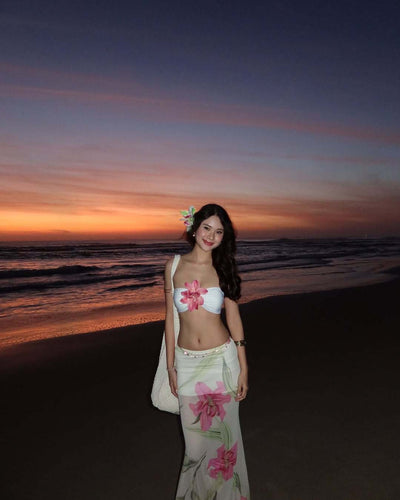 Woman in a white tube top with pink flower and a sheer floral maxi skirt, posing on a beach at sunset. - Image 8