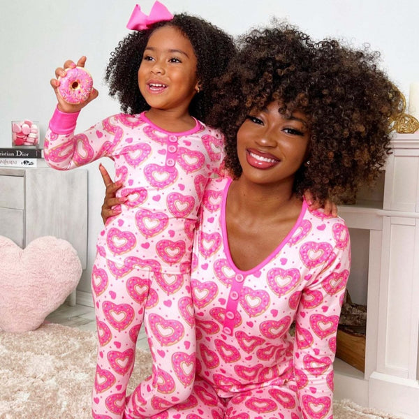 Mother and daughter in matching black and pink heart-print long-sleeve pajamas, smiling and holding small pink hearts.
