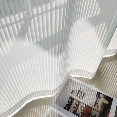 White vertical blinds with a pleated texture, allowing sunlight to filter through and cast shadows on an open magazine. - Image 4