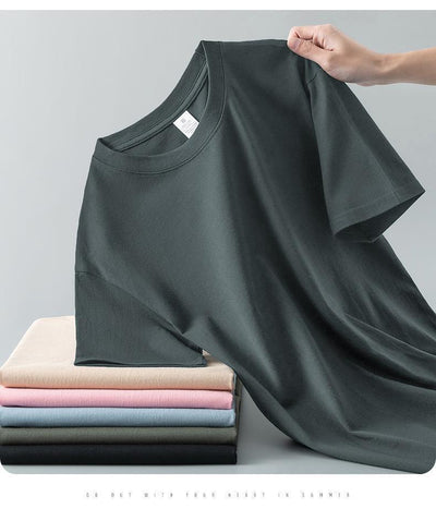 Light gray short-sleeve t-shirt with a round neck, held up to show its smooth fabric. A stack of folded t-shirts in various colors is visible in the background. - Image 29