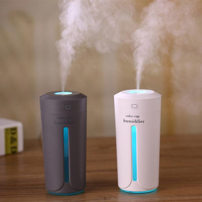 White mini USB ultrasonic humidifier with a green LED light strip, emitting mist, on a white surface.
