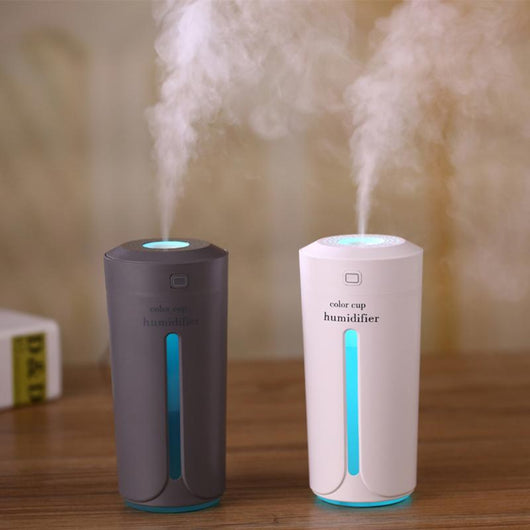 White mini USB ultrasonic humidifier with a green LED light strip, emitting mist, on a white surface.