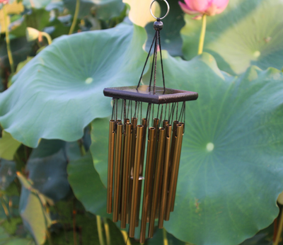 Bronze wind chime with multiple metal tubes hanging from a dark rectangular top, set against green lotus leaves. - Image 10