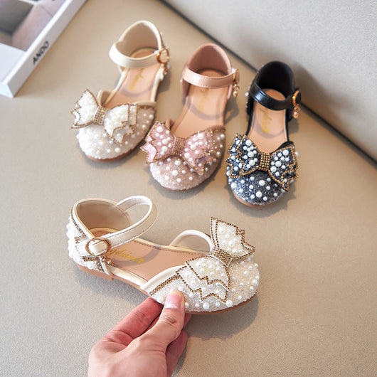 Beige girls' shallow mouth pumps with a large pink pearl bow, glitter, and an ankle strap with a gold buckle.