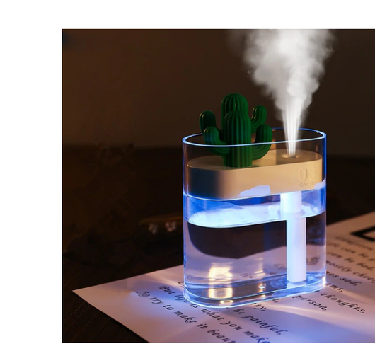 Transparent portable humidifier with a green cactus design on top, emitting mist on a wooden desk next to a laptop.