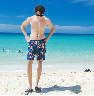 Man wearing navy blue swim shorts with a vibrant orange and green floral pattern, standing on a sunny beach. - Image 9
