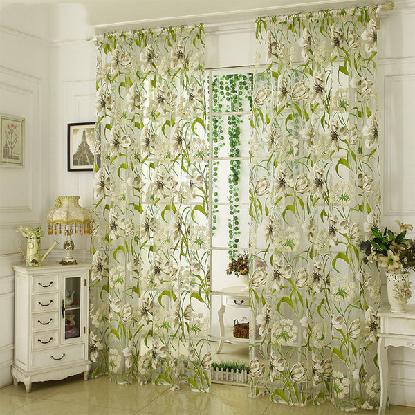 White sheer curtains with a green and beige floral pattern, hanging on a rod in a bright room.
