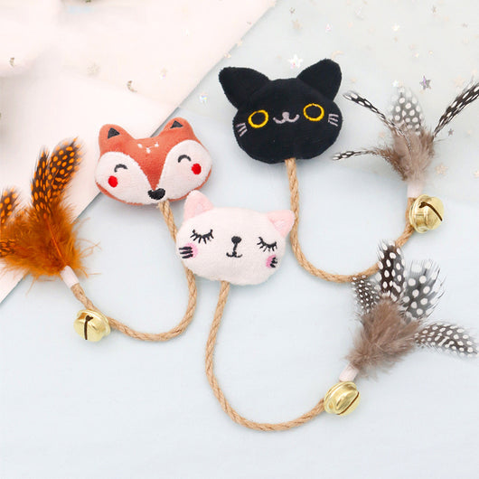 White cat head plush teaser toy with hemp rope, gold bell, and natural spotted feathers on pink packaging.