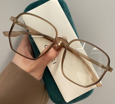 Mocha brown irregular frame glasses with clear lenses, held over a dark green velvet glasses case. - Image 10