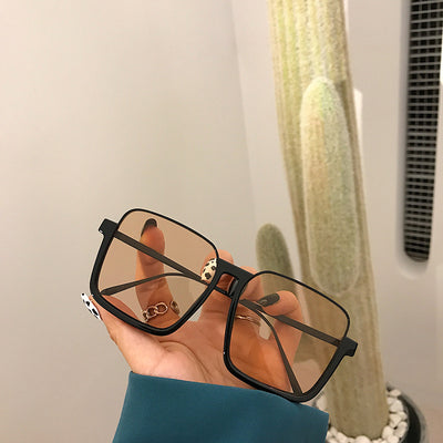 Retro square sunglasses with black frames and brown lenses held by a hand with patterned nails and a gold ring.