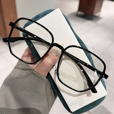 Mocha brown irregular frame glasses with clear lenses, held over a dark green velvet glasses case. - Image 4