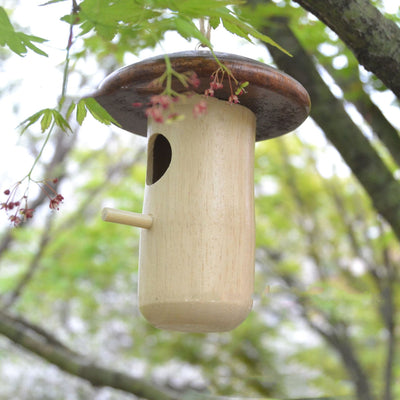 Wooden mushroom-shaped hummingbird feeder with a dark brown cap and light wood body, featuring an entrance hole and perch, held in a hand outdoors. - Image 2
