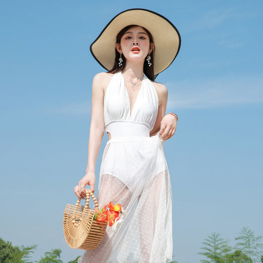 Woman in a white backless halter swimsuit with a sheer polka dot cover-up skirt, straw hat, and bamboo bag.
