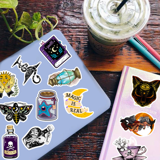 A hand holds a gothic-themed sticker featuring a raven, skull, and crystals, with a pile of 50 assorted waterproof stickers in the background.
