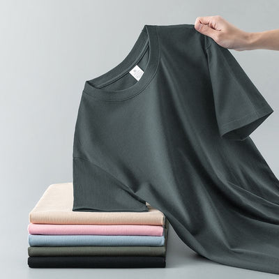 Light gray short-sleeve t-shirt with a round neck, held up to show its smooth fabric. A stack of folded t-shirts in various colors is visible in the background. - Image 4