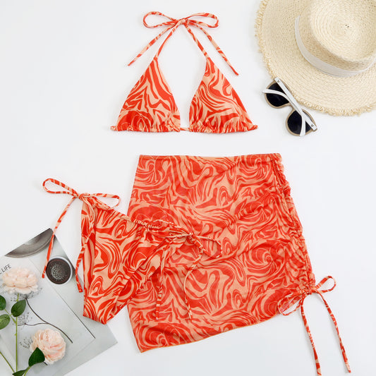 Women's three-piece swimsuit in red and orange abstract swirl print, featuring a halter bikini top, tie-side bottoms, and a ruched mini skirt.