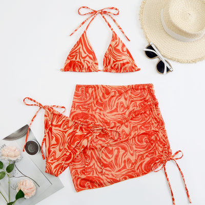 Women's three-piece swimsuit in red and orange abstract swirl print, featuring a halter bikini top, tie-side bottoms, and a ruched mini skirt.