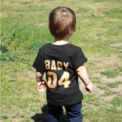 Child's black short-sleeved t-shirt with 'BABY 04' in gold print on the back, worn by a toddler in grass. - Image 6