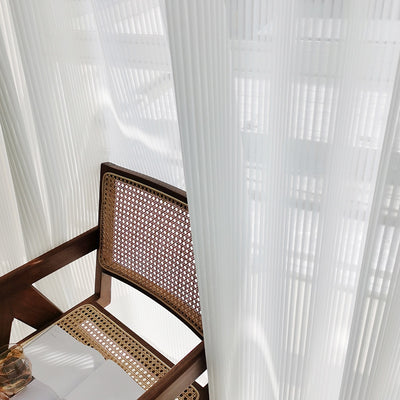 White vertical blinds with a pleated texture, allowing sunlight to filter through and cast shadows on an open magazine. - Image 3