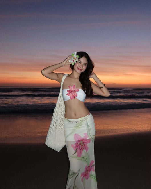 Woman in a white tube top with pink flower and a sheer floral maxi skirt, posing on a beach at sunset. - Image 12