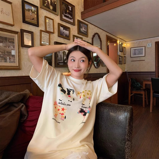 White loose-fit short-sleeve top with colorful retro cartoon cow print, modeled by a woman with plaid pants. - Image 2