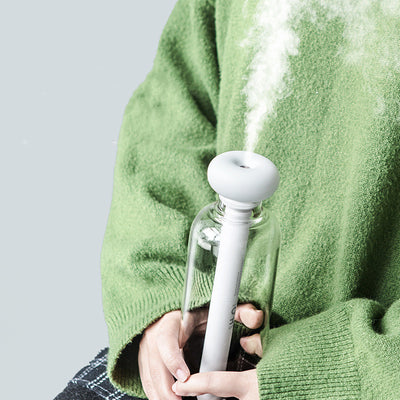 Clear bottle-shaped USB humidifier with a white donut-style top, emitting a stream of cool mist on a wooden table. - Image 5